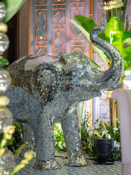 A mirrored elephant statue adds a touch of modern whimsy to the traditional 'Kel Baradari' wedding decor. It reflects the surrounding greenery and light, creating a dynamic visual element.