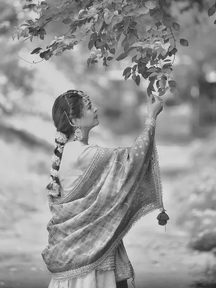 A beautiful black and white portrait that emphasizes the texture and form of the traditional outfit. Her interaction with the leaves adds a touch of gentle curiosity.