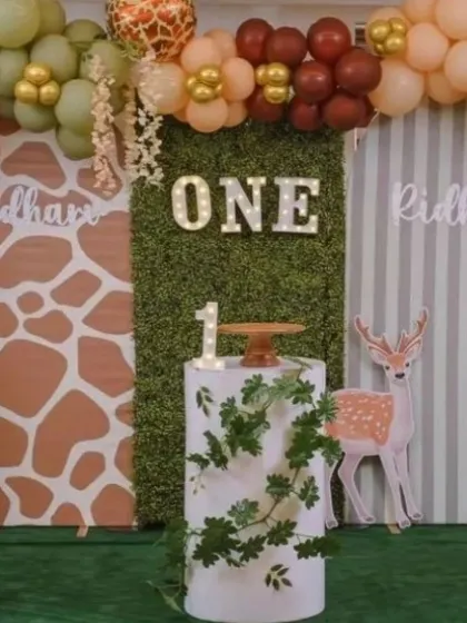 A rustic woodland-themed first birthday with a light-up "ONE" sign against a greenery panel. The decor uses earthy balloon colors and a giraffe-print backdrop for a charming effect.