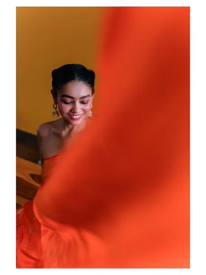 Using creative framing, this artistic shot captures a smiling model in an orange saree, partially obscured by a sweep of the fabric. It creates a sense of mystery and focuses on the emotion of the moment.