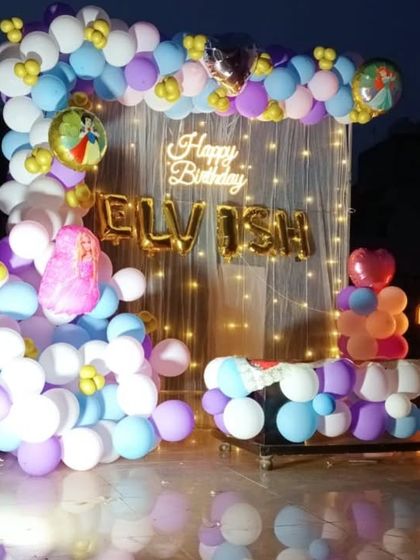 A first birthday party with a pastel balloon arch, fairy lights, and character foil balloons.
