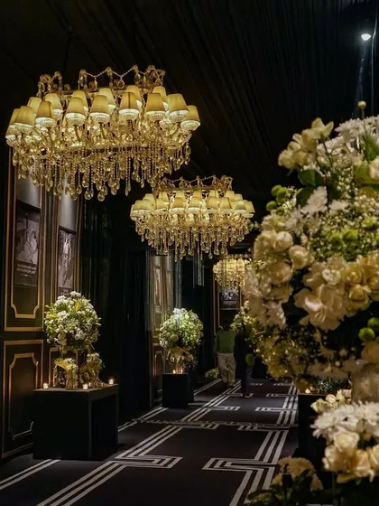 The grand entrance to the anniversary party, featuring a dramatic black-and-gold color scheme, massive chandeliers, and abundant white floral arrangements, setting a tone of timeless elegance.