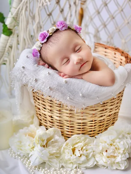 A sleeping baby rests its head on its hands in a wicker basket, wearing a delicate floral crown. This pose highlights the baby's peaceful expression and tiny features.