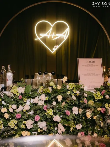 The bar at the "Big Bow Affair," featuring a custom neon sign with the couple's initials, a lush floral front, and a well-stocked setup.