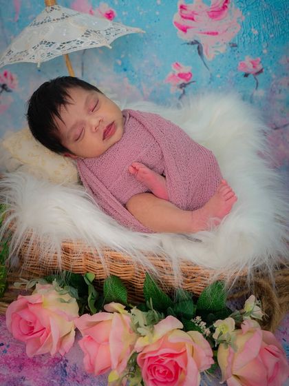 A wider shot showing the newborn girl in a basket with a tiny lace umbrella. The combination of textures like fur, flowers, and lace creates a beautiful, soft image.
