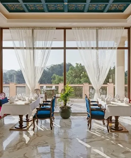 A sun-drenched dining area at IHCL Pilibhit-Haridwar, where floor-to-ceiling windows frame the lush outdoors. The vibrant pink and blue seating adds a pop of color to the serene, marble-floored space.