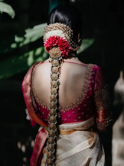A beautiful back shot of a bride in a pink and cream Kanchipuram saree. The blouse features a deep back with intricate gold embroidery, perfectly complementing her traditional braided hairstyle and temple jewelry.