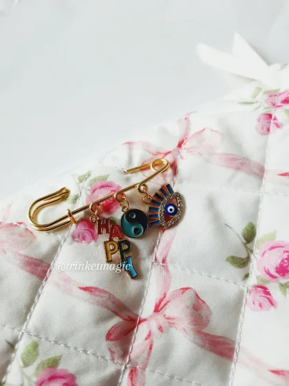 A happy and protective charm pin. This gold safety pin is adorned with 'HAPPY' letter charms, a yin-yang symbol, and an evil eye, perfect for spreading good vibes.