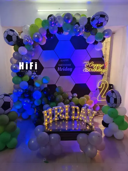 A unique football-themed birthday decoration for a young sports fan. The backdrop is designed like a soccer ball, framed by a green, black, and white balloon arch, complete with a neon sign and marquee letters.