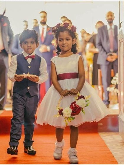 A beautiful flower girl walking down the aisle. Her classic white A-line dress with a simple maroon sash is a testament to timeless elegance and youthful grace.