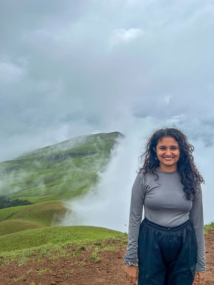 A trekker with beautiful curly hair smiles against the backdrop of the misty Netravathi peaks.