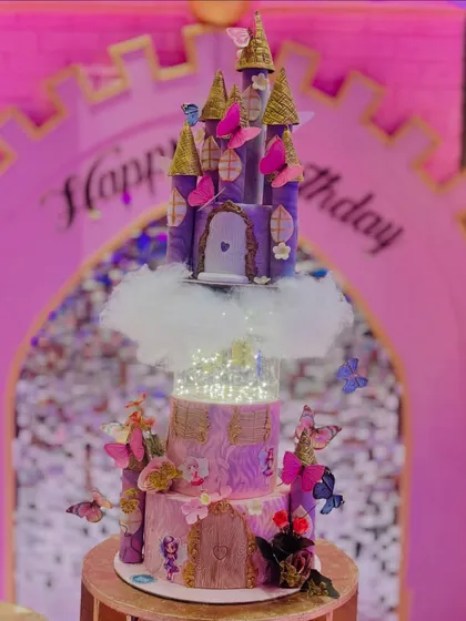 A majestic purple and pink rotating castle cake, set up for a royal birthday party. The middle tier is illuminated with fairy lights, and the entire structure is surrounded by edible clouds and butterflies.