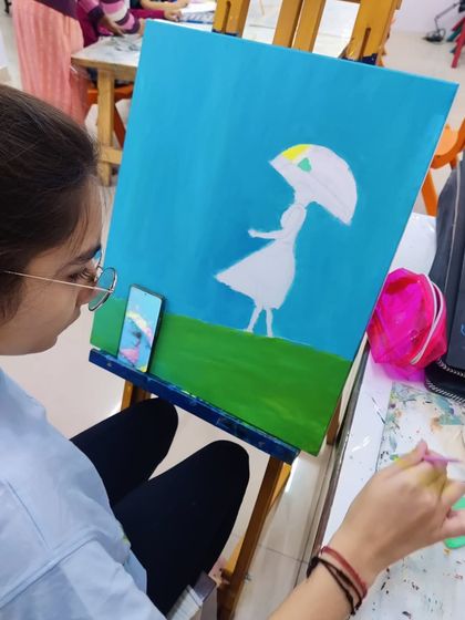 A student works on an imaginative painting of a girl with an umbrella in a field, using a palette knife and brush to create texture.