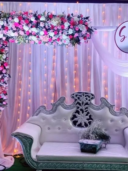 An elegant engagement stage with a white and silver carved sofa. The decor is dominated by a large L-shaped floral arrangement of pink and white roses, set against a backdrop of fairy lights and a custom monogram with the couple's initials.