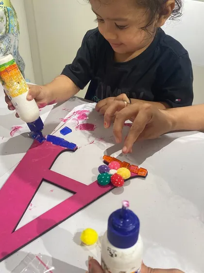 Even the littlest artists can join in the fun! This toddler is getting some help gluing colorful beads and mosaics onto her monogram. It's a great sensory activity for developing fine motor skills.