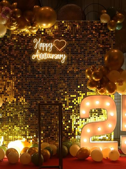 A gorgeous 25th-anniversary celebration featuring a gold shimmer wall, a 'Happy Anniversary' neon sign, large marquee numbers, and an elegant balloon arch in olive green, cream, and gold.
