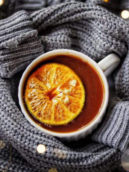 Happiness is warming your hands with a hot cup of tea. This top-down shot shows a cup of orange-infused tea nestled within the sleeves of a cozy gray sweater, surrounded by fairy lights.
