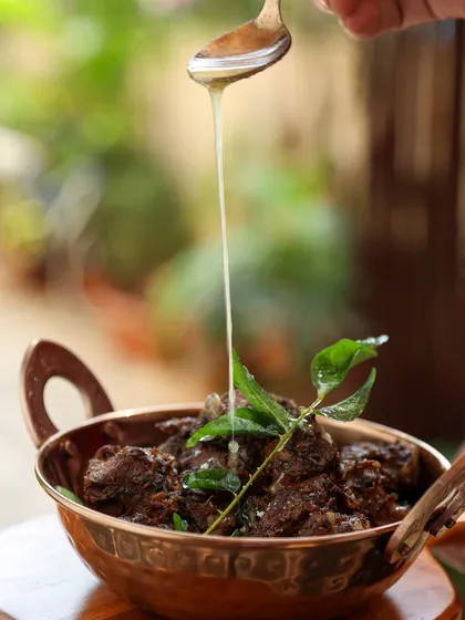 A slow, mesmerizing pour of golden ghee over mutton sukka. This action shot emphasizes richness and tradition.