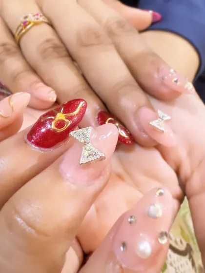 A vibrant and playful set combining red nails with gold art and sheer nails decorated with pearls, crystals, and a large silver rhinestone bow charm.