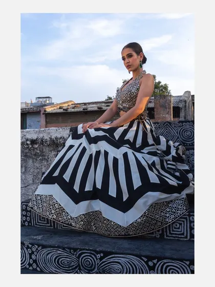 A full view of a model sitting gracefully in a black and white geometric-patterned lehenga. The composition highlights the volume and design of the skirt, making it ideal for a designer lookbook.