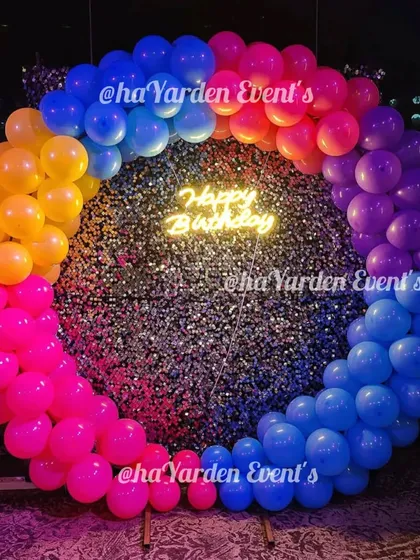 A vibrant, multi-color balloon ring set against a silver sequin wall. The 'Happy Birthday' neon sign in the center makes this a fun and eye-catching photo spot.
