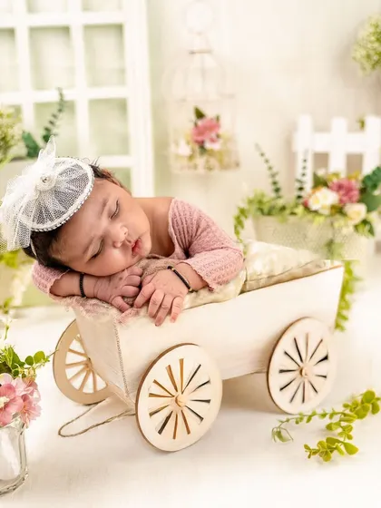 Dressed in soft pink, this baby girl sleeps peacefully in a charming little wooden cart. The surrounding floral arrangement adds a touch of garden magic to this newborn posing idea.