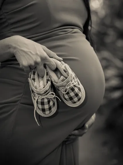 A close-up, black and white shot focusing on a pair of tiny plaid baby shoes held against the baby bump. A classic and artistic detail shot.