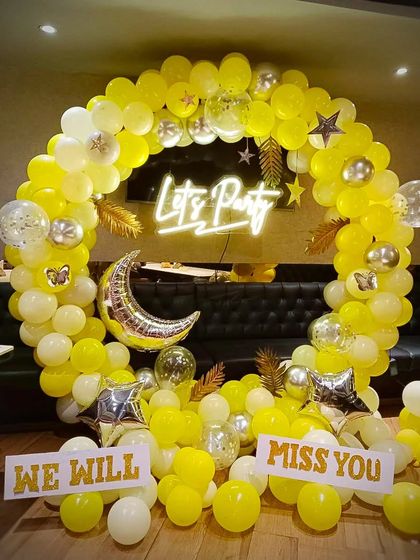 A bright and cheerful setup for a farewell party. The yellow and white balloon ring, "Let's Party" neon sign, and "WE WILL MISS YOU" signs create a positive and celebratory atmosphere for saying goodbye to a colleague.