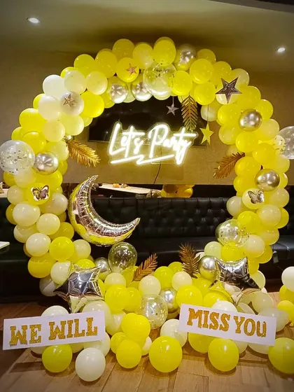 A bright and cheerful setup for a farewell party. The yellow and white balloon ring, "Let's Party" neon sign, and "WE WILL MISS YOU" signs create a positive and celebratory atmosphere for saying goodbye to a colleague.