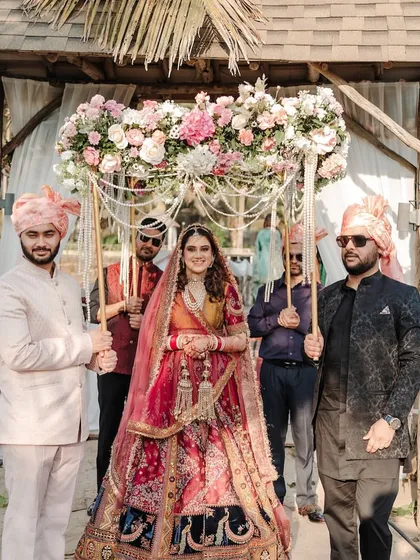 A beautiful bridal entry under a 'phoolon ki chadar' (floral canopy) at a beach wedding. The rustic wooden structure complements the natural, sandy surroundings perfectly.