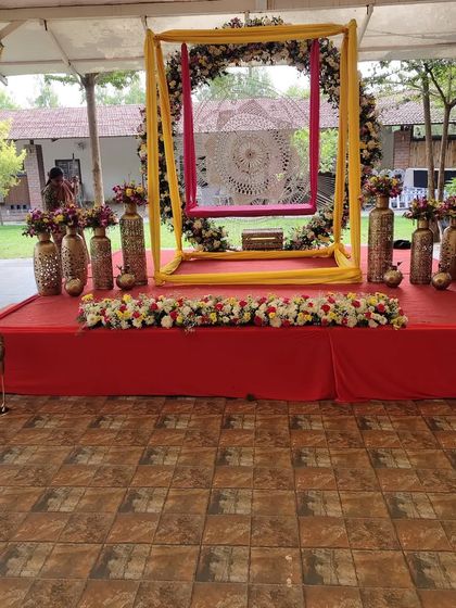 A lovely Mehendi setup featuring a floral swing on a red stage, surrounded by brass vases filled with flowers.