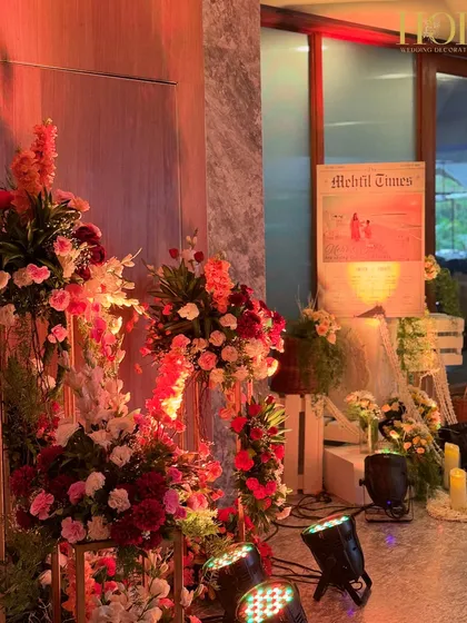 A rustic and charming welcome setup featuring a personalized "Mehfil Times" newspaper sign, surrounded by floral arrangements in wicker baskets and warm candle lights.