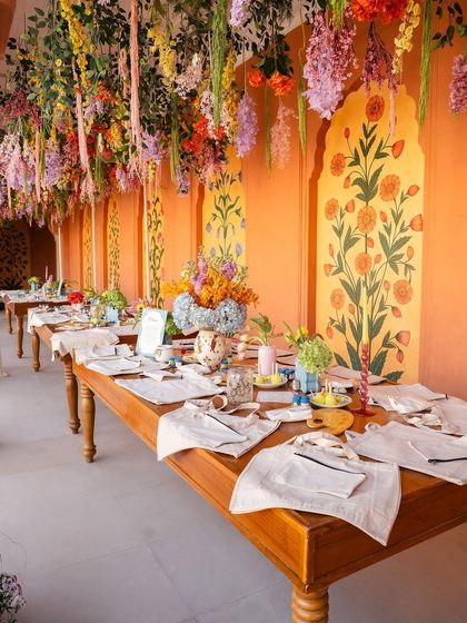 The setup for our "Cultural Canvas" art activity. Long wooden tables were placed under a ceiling of hanging colorful flowers, with art supplies neatly arranged at each place setting for guests to get creative.