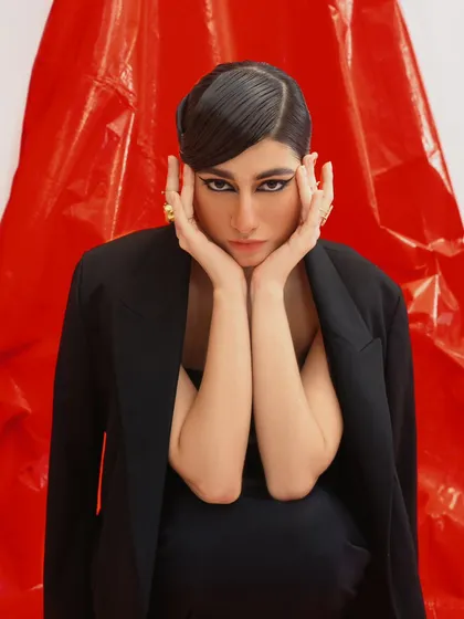 A dramatic beauty shot from a modern fashion editorial. The sharp, graphic eyeliner and the model's pose are highlighted against a vibrant red backdrop.