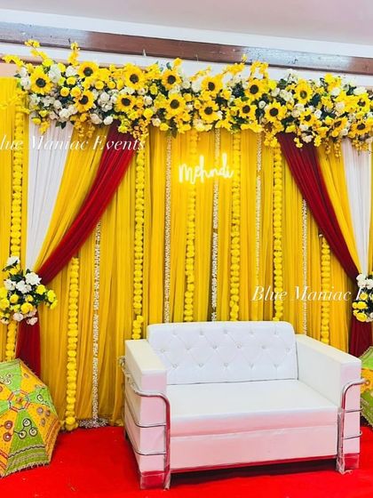 A classic Haldi or Mehendi setup with a bright yellow backdrop, marigold garlands, and a pop of red and white draping. The 'Mehendi' neon sign and traditional umbrellas add to the festive charm.