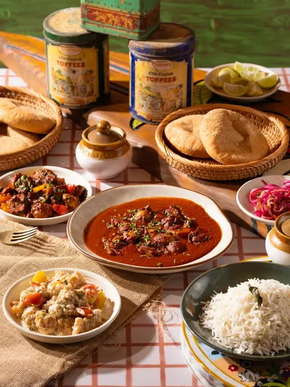 A spread of Goan food, including a rich curry, rice, and bread. The checkered tablecloth gives it a homey, rustic cafe vibe.