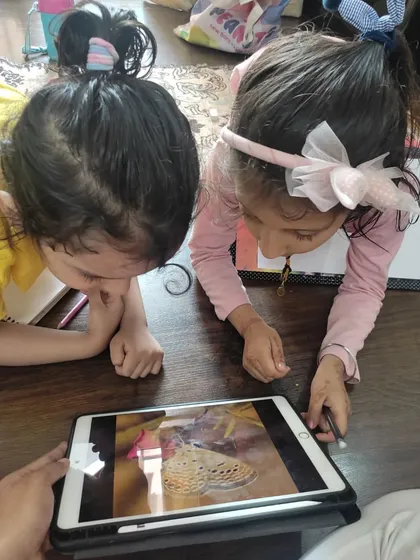 Curiosity is the first step in art. Before drawing, these two girls are closely observing a picture of a butterfly on a tablet, learning about its patterns and colors to inspire their own creations.