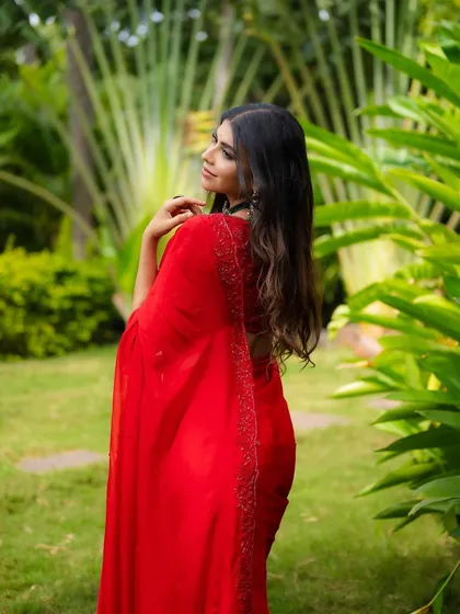 A portrait of a model in a vibrant red saree, seen from the back. The focus is on the beautiful fabric and her flowing hair against the green background.