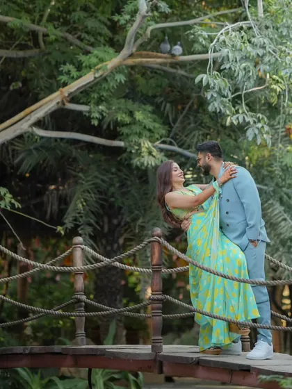 A playful and romantic moment on a rustic wooden bridge. The bride, in a vibrant turquoise and yellow saree, shares a joyful embrace with her partner, who is dressed in a stylish light blue suit.