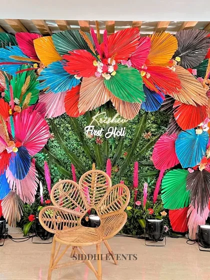 For a 'First Holi' celebration, we created this explosion of color. A lush green wall serves as the base for vibrant, multi-colored dried palm leaves, creating a joyful and energetic photo backdrop. The natural rattan peacock chair adds a touch of bohemian chic to the setup.