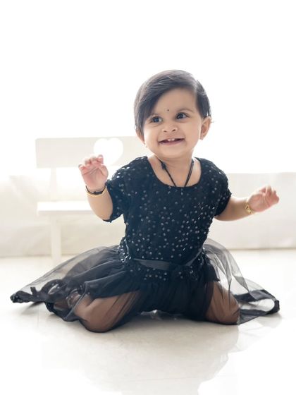 A happy toddler in a black party dress, kneeling and smiling. The simple white background keeps the focus on her.