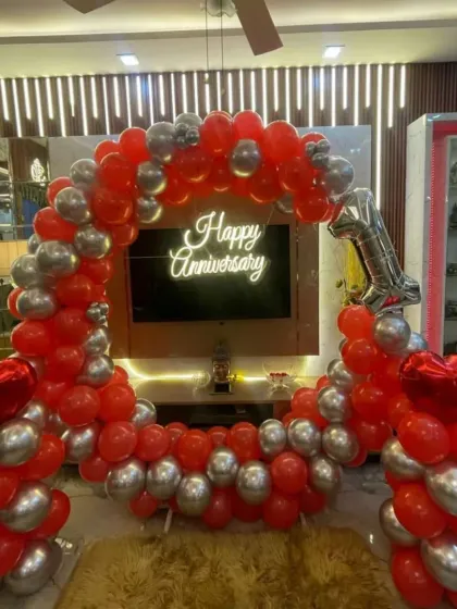 This vibrant first-anniversary decoration creates a festive mood right in your living room. A full balloon arch in bold red and metallic silver frames the TV area, highlighted by a "Happy Anniversary" neon sign and a number "1" foil balloon.