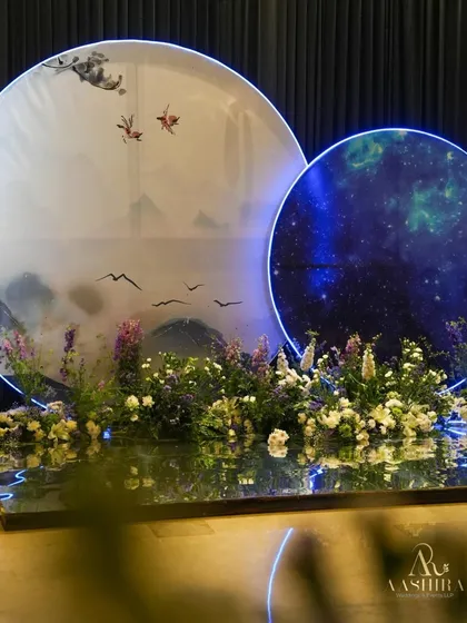 A unique photo corner with two circular backdrops, one depicting misty mountains and the other a glowing galaxy, set over a reflective floor with a bed of flowers.