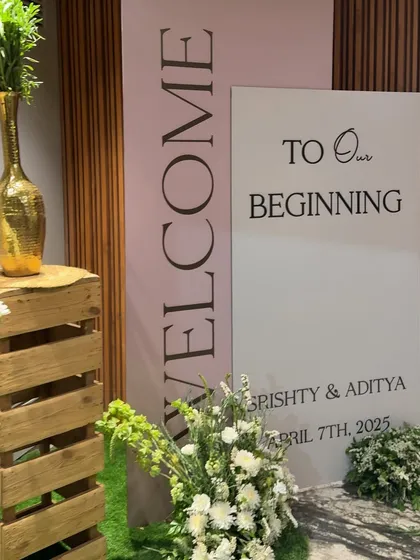 A modern and chic welcome for Srishty and Aditya's beginning. This setup features clean typography on dual panels, rustic wooden crates, and fresh white and green floral arrangements.