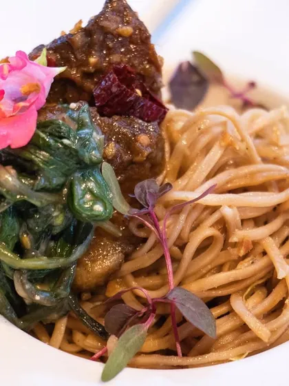 An artfully plated bowl of noodles with a skewer of marinated meat and a side of seasoned spinach, garnished with microgreens and an edible flower.