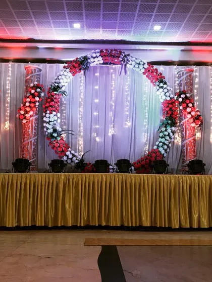 A popular choice for modern receptions, this stage features a large circular floral wreath as the central backdrop element. The setup is framed by lit panels, creating a bright and festive look.