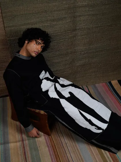 An artistic menswear shot for a campaign, featuring a model in a long tunic with a bold, abstract white print. The earthy, textured background complements the avant-garde design of the garment.
