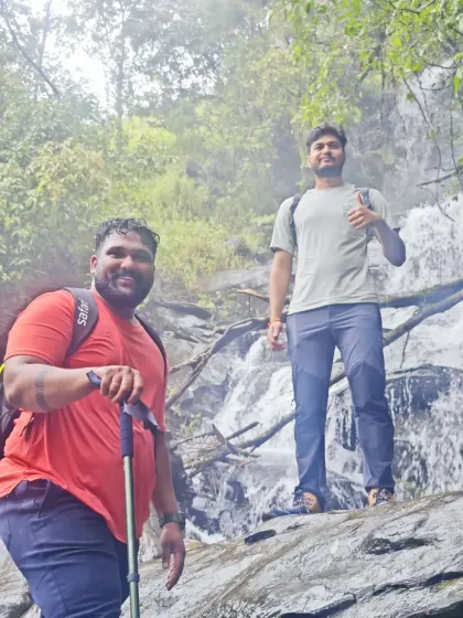Two trekkers posing in front of a waterfall, fully equipped with backpacks and trekking poles.