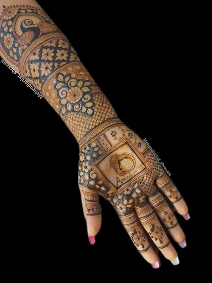 A single hand showing a mix of traditional and custom elements. The palm features a ring illustration with the name 'Anand', while the forearm has classic peacock and floral patterns.