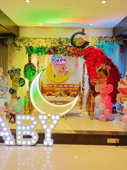 A grand baby shower stage with a peacock floral arrangement, a lighted moon, and 'BABY' marquee letters. The setup includes 'It's a Boy' and 'It's a Girl' cutouts.